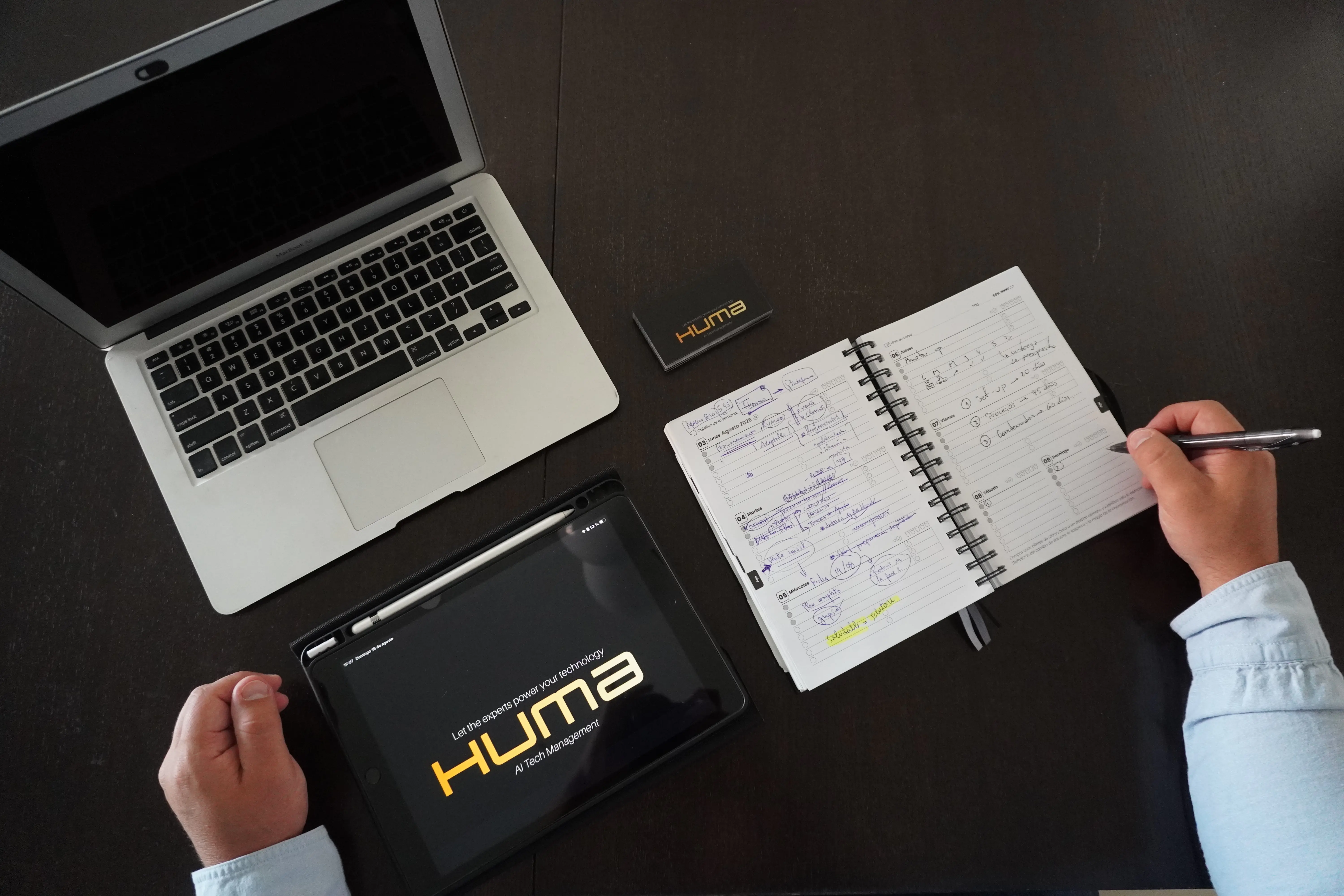 Escritorio de trabajo visto desde arriba con laptop, tablet mostrando el logo de Huma AI Tech Management y una agenda con planificación estratégica escrita a mano